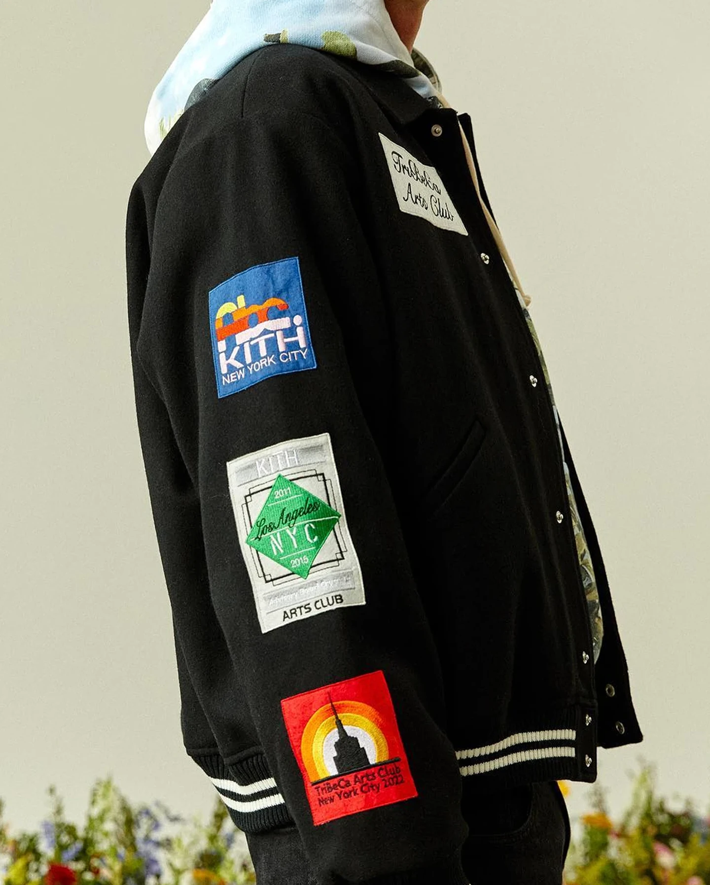 Model wearing a black KITH varsity jacket covered with colorful embroidered patches.