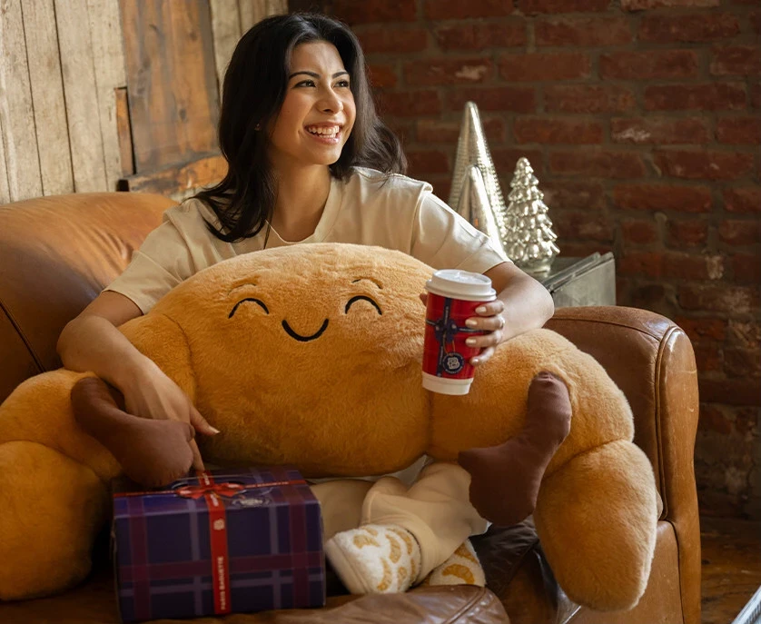 Woman seated for the holidays with an oversized smiling croissant plush, branded coffee cup, and gift box.