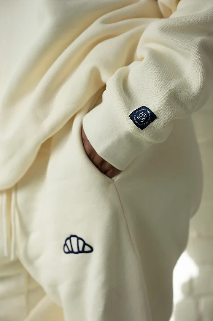 Close-up of a cream crewneck with a navy embroidered croissant and a Paris Baguette cuff label.