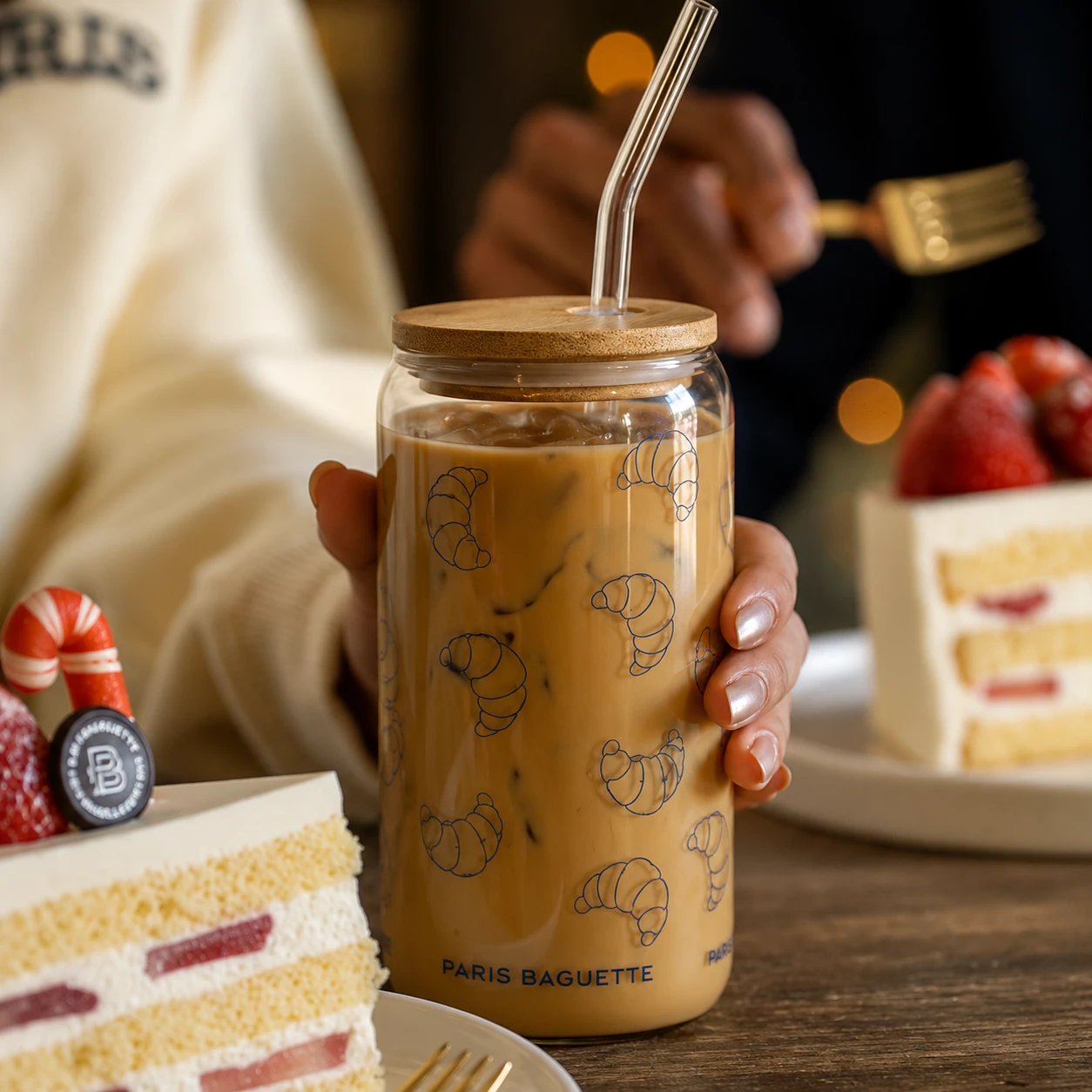 Glass tumbler with a bamboo lid, clear straw, and repeating blue croissant illustrations, filled with iced coffee.