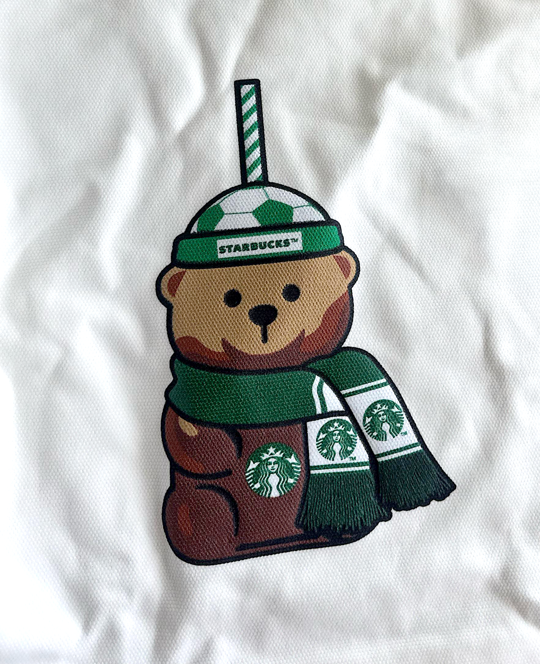 Close-up of an embroidered Starbucks Bearista character wearing a green scarf on white fabric.