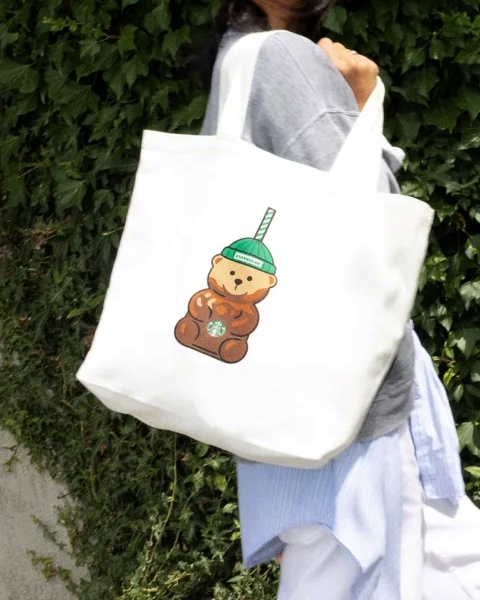 Woman carrying a cream Starbucks tote printed with the Bearista character.